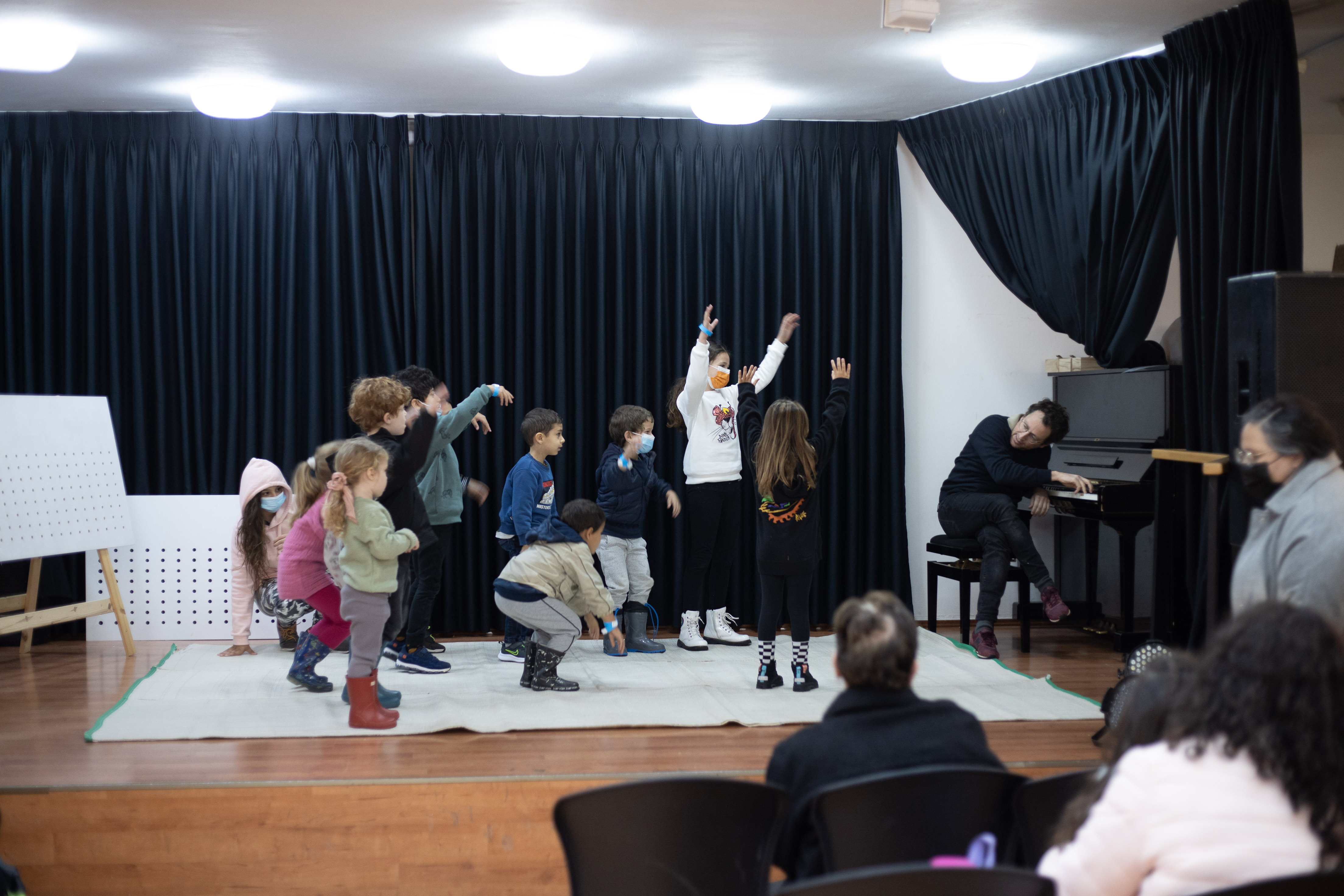 צילום מתוך סדנת ההלחנה בבית מאניה ובו ההורים הישובים בקהל מביטים בילדיהם על הבמה מגיבים למוסיקה אותה מנגן המדריך על הפסנתר